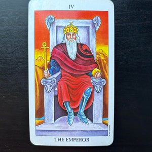 Puede incluir: La carta del tarot El Emperador presenta una figura con una t&uacute;nica roja, barba blanca y corona, sentada en un trono con cabezas de carnero. La carta est&aacute; numerada IV y tiene un fondo rojo, naranja y amarillo.