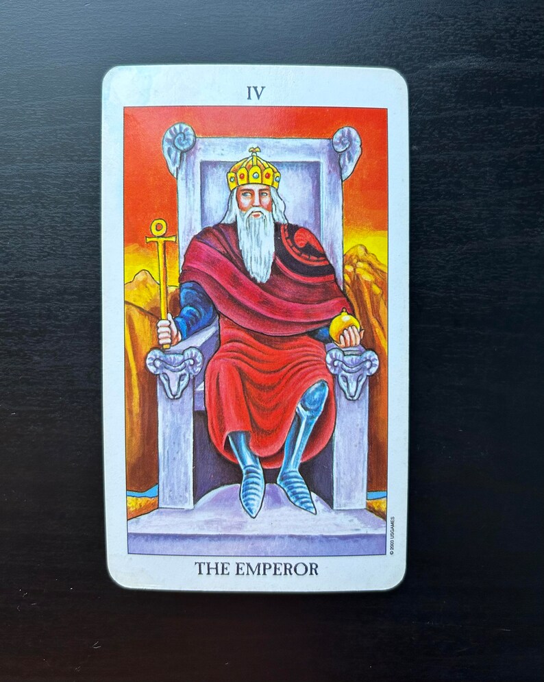 Puede incluir: La carta del tarot El Emperador presenta una figura con una t&uacute;nica roja, barba blanca y corona, sentada en un trono con cabezas de carnero. La carta est&aacute; numerada IV y tiene un fondo rojo, naranja y amarillo.