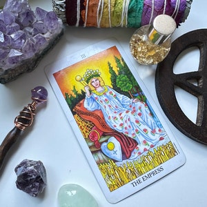 Puede incluir: Una lectura de cartas del tarot con la carta de la Emperatriz, un c&uacute;mulo de cristal de amatista p&uacute;rpura, un palo de salvia envuelto en p&eacute;talos de flores de colores, una varita de madera con un cristal morado, una peque&ntilde;a botella de purpurina dorada y un signo de paz de madera.