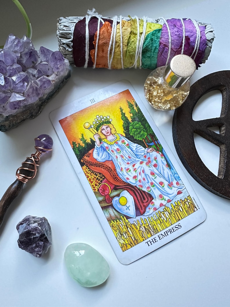 Puede incluir: Una lectura de cartas del tarot con la carta de la Emperatriz, un c&uacute;mulo de cristal de amatista p&uacute;rpura, un palo de salvia envuelto en p&eacute;talos de flores de colores, una varita de madera con un cristal morado, una peque&ntilde;a botella de purpurina dorada y un signo de paz de madera.