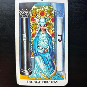Puede incluir: La carta del tarot La Sacerdotisa Mayor muestra a una mujer con una t&uacute;nica azul sentada en un trono entre dos pilares. Lleva una corona con una luna creciente y sostiene un rollo en el que se lee "TORA". La carta est&aacute; situada sobre un fondo de jard&iacute;n con flores y un cielo azul.