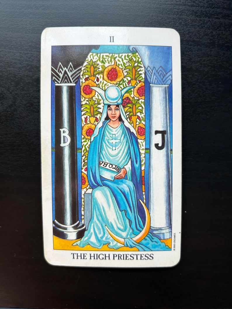 Puede incluir: La carta del tarot La Sacerdotisa Mayor muestra a una mujer con una t&uacute;nica azul sentada en un trono entre dos pilares. Lleva una corona con una luna creciente y sostiene un rollo en el que se lee "TORA". La carta est&aacute; situada sobre un fondo de jard&iacute;n con flores y un cielo azul.