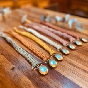 Peut inclure: Plusieurs attaches-sucettes en macramé de couleurs variées, beige, jaune et rose. Chaque attache présente un motif tissé et un clip en métal argenté avec une pièce en plastique ronde, transparente et irisée.