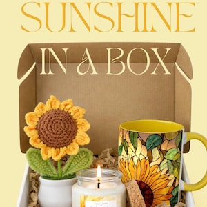 以下が含まれることがあります： 「SUNSHINE IN A BOX」と書かれたギフトボックスには、かぎ針編みのヒマワリ、ヒマワリのデザインの黄色いマグカップ、火のついたキャンドルが入っています。ボックスは装飾用の紙で満たされています。