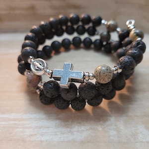 May include: A black lava rock and brown stone beaded bracelet with a silver cross and a silver charm. The bracelet is wrapped around a wooden surface.