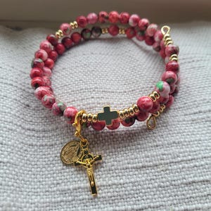 Puede incluir: Un brazalete de cuentas rojas y blancas con un dije de cruz dorada y un dije de medalla dorada. El brazalete está hecho de pequeñas cuentas redondas con un patrón floral.
