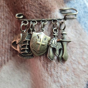 May include: A gold-toned metal brooch with five charms: a shield with a cross, a helmet, a sword, and two other charms. The brooch is on a brown fuzzy surface.