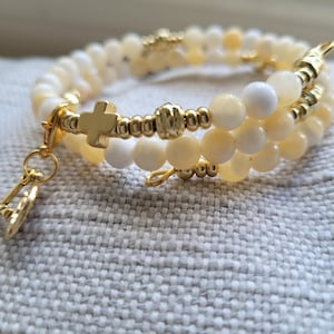 May include: A double-strand bracelet with white and yellow beads and gold accents. The bracelet features a small gold cross charm and a key charm.
