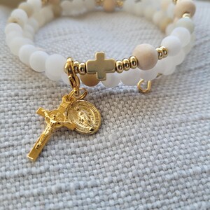 May include: A white and wood bead bracelet with a gold cross charm and a gold pendant featuring the Virgin Mary.
