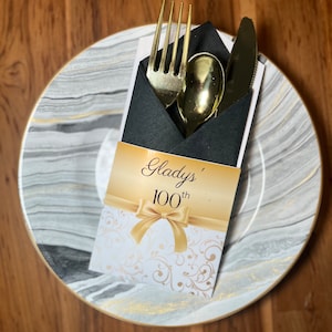 May include: A black napkin with gold cutlery and a white card with gold lettering that reads "Gladys' 100th" on a marble plate.