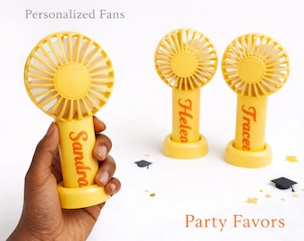 May include: Yellow personalized handheld fans with names in orange script. One fan is held by a hand, while two others stand upright. The text "Personalized Fans" and "Party Favors" are also visible. Graduation cap and star confetti are scattered around.