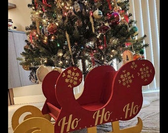 以下が含まれることがあります： 赤い木製のそりで、金色のアクセントと「Ho Ho Ho」の文字が側面に書かれています。そりは雪の結晶で飾られており、クリスマスツリーの前に置かれています。