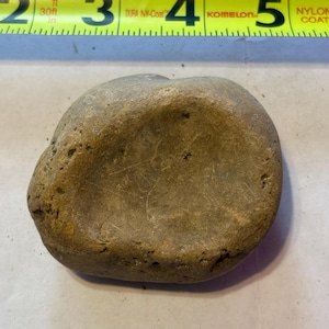 Puede incluir: Una piedra lisa, redondeada, de color marrón claro, con una depresión cóncava. La piedra mide aproximadamente 10 cm de largo, medida por una cinta métrica amarilla en el fondo. La cinta métrica tiene marcas y texto en negro.