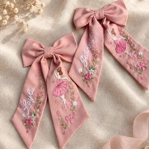 Op de afbeelding: Twee oudroze stoffen haarstrikken met geborduurde ballerinafiguren in roze tutu's, bloemenkransen en de namen Layla en Jeilany gestikt in wit garen. De strikken hebben lange, puntige uiteinden en zijn gemaakt van zacht, mat textielmateriaal.