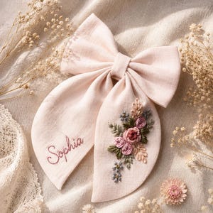 Pode incluir: Um laço de cabelo em tecido rosa pálido com o nome Sophia bordado numa das pontas e um design floral detalhado com fios rosa, verdes e azuis na outra ponta. O laço tem uma forma estruturada com duas pontas longas e arredondadas.