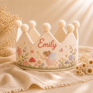 Puede incluir: Una corona de fieltro color crema con patrones bordados de flores, setas y una figura de hada. El nombre Emily está bordado en hilo rosa en la parte frontal. La corona está rematada con pequeños pompones crema en cada una de sus seis puntas.