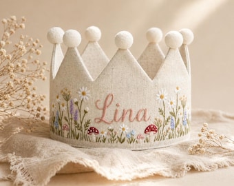 Könnte beinhalten: Eine beige Stoffkrone mit floraler Stickerei, die Gänseblümchen, Lavendel und Pilze zeigt. Der Name "Lina" ist rosa gestickt. Die Krone hat weiße Pompons auf jeder Spitze.