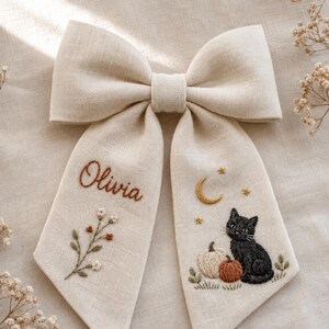 Peut inclure: Un nœud pour cheveux en lin beige avec des broderies faites à la main. Le pan gauche porte le prénom Olivia en lettres cursives brunes au-dessus d'une tige fleurie. Le pan droit montre un chat noir près de deux citrouilles sous un croissant de lune.