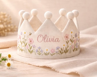 Könnte beinhalten: Eine cremefarbene Stoffkrone mit gestickten Blumen in Rosa, Lila und Weiß und dem Namen "Olivia". Die Krone hat weiße Pompons und eine Biene. Ein bezauberndes Accessoire.