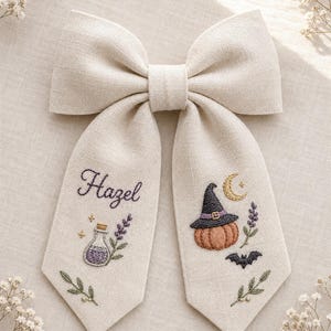 May include: A beige linen hair bow featuring embroidered designs on its two long tails. The left tail displays the name Hazel, a purple potion bottle, and lavender sprigs. The right tail shows a pumpkin wearing a witch hat, a crescent moon, a bat, and lavender.