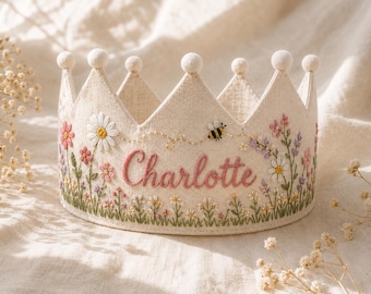 May include: A cream-colored fabric crown featuring embroidered wildflowers, a small bee, and the name Charlotte stitched in pink thread. The crown has six points, each topped with a small white pom-pom, and is decorated with green stems.