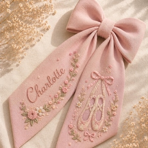 Può includere: Un fiocco per capelli in tessuto rosa polvere con due nastri lunghi. Un nastro è ricamato con il nome Charlotte in corsivo circondato da motivi floreali. L'altro nastro mostra un paio di scarpette da danza classica ricamate con nastri.