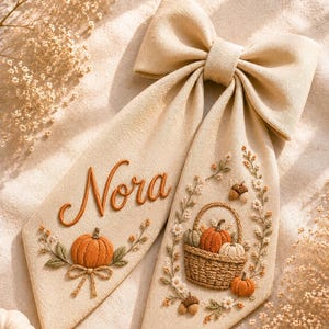 Puede incluir: Un lazo para el pelo de tela beige con detalles bordados. Un extremo muestra el nombre Nora en letra cursiva naranja sobre una calabaza bordada. El otro extremo presenta una cesta llena de calabazas, rodeada de motivos florales y bellotas.