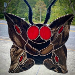 May include: Stained glass moth with large red eyes and brown wings. The moth has black antennae and red accents. The glass is framed with a silver metal. The artwork is hanging from a chain.