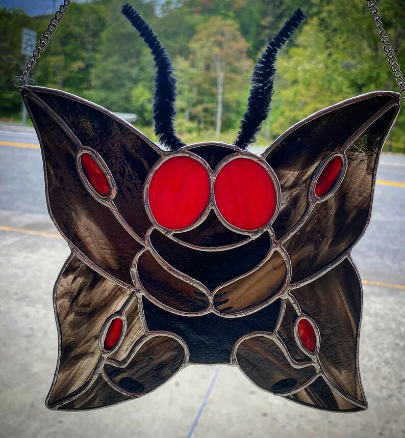 May include: Stained glass moth with large red eyes and brown wings. The moth has black antennae and red accents. The glass is framed with a silver metal. The artwork is hanging from a chain.