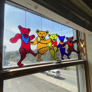 Pode incluir: Suncatchers de vidro manchado de ursos dançantes em vermelho, amarelo, verde, azul e castanho, cada um com um lenço colorido. Os ursos estão pendurados em frente a uma janela, com vista para uma estrada e árvores.