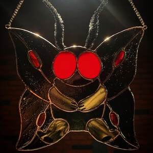 May include: Stained glass moth with large red eyes, black wings, and yellow and red accents. The artwork is suspended by a delicate chain, showcasing a unique, handcrafted design. The moth's body and wings are detailed with intricate glasswork.