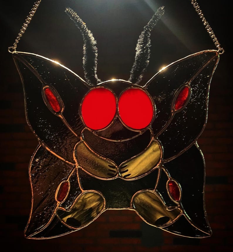 May include: Stained glass moth with large red eyes, black wings, and yellow and red accents. The artwork is suspended by a delicate chain, showcasing a unique, handcrafted design. The moth's body and wings are detailed with intricate glasswork.