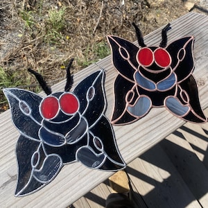 May include: Two stained glass butterfly ornaments. Each butterfly features a black body with red eyes and gray and blue wing accents. The butterflies have black pipe cleaner antennae and are displayed on a wooden surface.