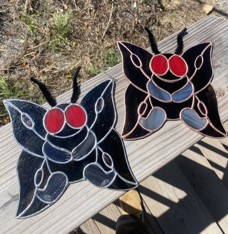 May include: Two stained glass butterfly ornaments. Each butterfly features a black body with red eyes and gray and blue wing accents. The butterflies have black pipe cleaner antennae and are displayed on a wooden surface.