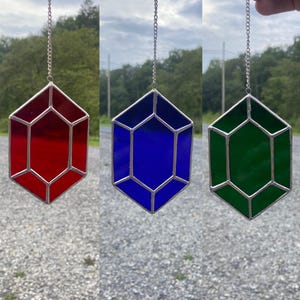 Op de afbeelding: Drie geometrische hangers van glas in lood in rood, blauw en groen, elk met een zilverkleurig metalen frame en ketting. De hangers hangen tegen een vage buitenachtergrond.