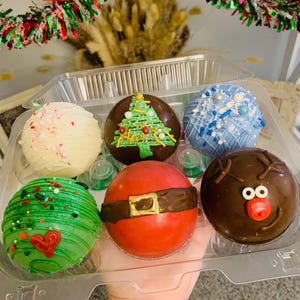 May include: A clear plastic container holding six festive, decorated chocolate spheres. Designs include a Christmas tree, Santa Claus, a reindeer, and snowflake patterns. The spheres are in shades of red, green, brown, white, and blue, perfect for holiday treats.