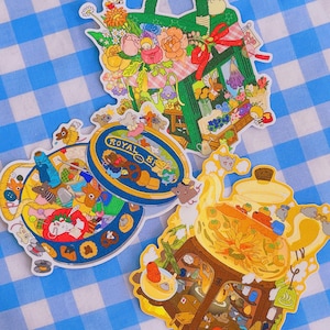 May include: Three colourful illustrated stickers featuring a teacup, a teapot, and a basket of flowers. Each sticker is filled with intricate details and whimsical characters. The stickers are on a blue and white checked background.