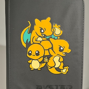 May include: A black zippered binder with a textured surface featuring a graphic of three orange dragon-like creatures. The word BYSTER is embossed in black at the bottom. The creatures are depicted in different poses with teal wing accents.