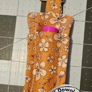 May include: A floral print lip balm holder in shades of orange and cream, with a pink lip balm peeking out. The holder has a metal clasp and a small loop. The word "Pattern" is in a white rectangle.