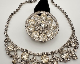Puede incluir: Un conjunto vintage de collar y broche de pedrería plateada. El collar presenta una delicada cadena con una serie de brillantes pedrería transparente. El broche es un diseño grande y redondo con un grupo de pedrería transparente. La joyería se exhibe sobre un soporte negro.