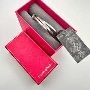 Puede incluir: Una pulsera plateada descansa en una caja rosa brillante con un interior gris. La caja está abierta, revelando la pulsera y una etiqueta con el nombre "Buckingham". La tapa de la caja también es visible, con el nombre "Buckingham" impreso.