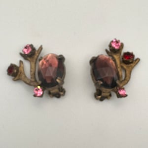 May include: A pair of antique earrings with a gold tone finish. Each earring features a dark purple faceted stone surrounded by a cluster of pink and red rhinestones. The earrings are shaped like a stylized branch with a cluster of leaves.