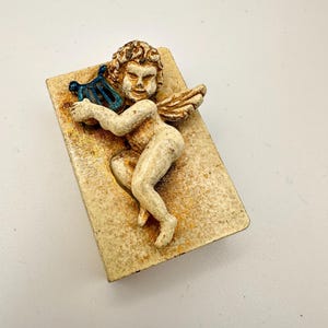 May include: A vintage-style figurine of a cherub playing a harp. The cherub is cream-colored with a weathered appearance, holding a blue harp. The figurine rests on a rectangular base with a textured, aged finish.