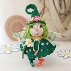 Puede incluir: Una muñeca de hada de ganchillo hecha a mano con un gorro puntiagudo verde adornado con flores. La muñeca tiene cabello verde, una nariz rosada y un vestido verde con detalles de flores blancas. Tiene pequeñas alas y zapatos naranjas y verdes. Una margarita está adjunta.