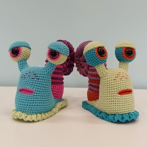 May include: Two crocheted snail toys, one blue with pink accents and the other green with orange accents. Both have large, black eyes and a crocheted shell.