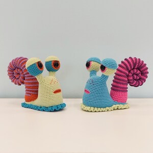 Könnte beinhalten: Zwei gehäkelte Schnecken-Plüschtiere. Eine ist blau mit rosa und weißen Akzenten, die andere ist gelb mit orange und blauen Akzenten.