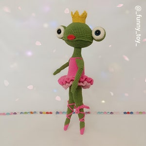 May include: A handmade green crochet frog doll wearing a pink tutu, a pink bodice, and a gold crown. The frog has large, expressive eyes and is posed as a ballerina. The doll's ballet slippers are pink and tied with pink ribbons.