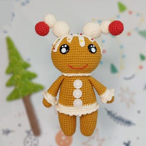 May include: A crocheted gingerbread person toy with white icing, red and white candy accents, and white buttons. The toy has a smiling face with black eyes and a red mouth.