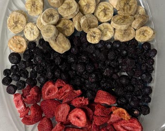 Puede incluir: Una bandeja de servir transparente con bordes festoneados contiene tres secciones distintas de fruta liofilizada: una capa superior de rodajas de plátano beige, una capa media de arándanos morados oscuros y una capa inferior de fresas rojas en rodajas.