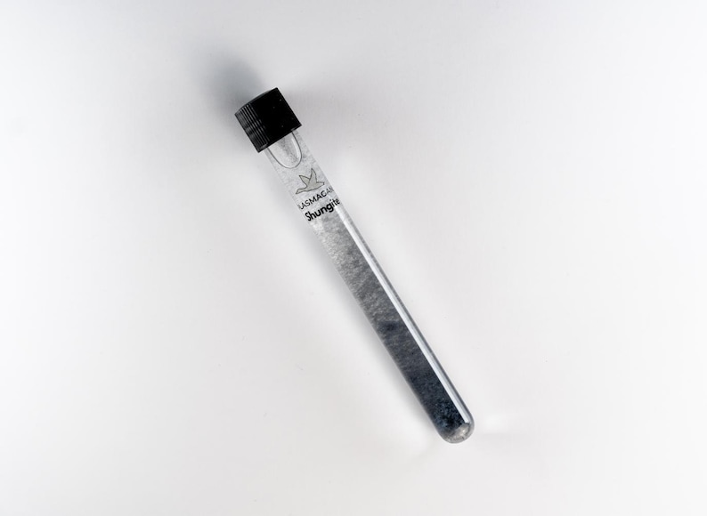 May include: A clear glass test tube with a black cap, filled with a dark grey substance. The tube has the text "LASMACAN Shungite" printed on it. The tube is angled against a white background.